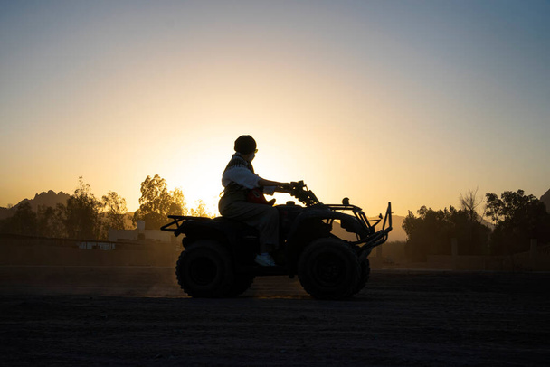 La silueta de una turista monta una quad bike sobre el fondo de una puesta de sol brillante. Noche de entretenimiento extremo en el desierto de Egipto. - Foto, imagen