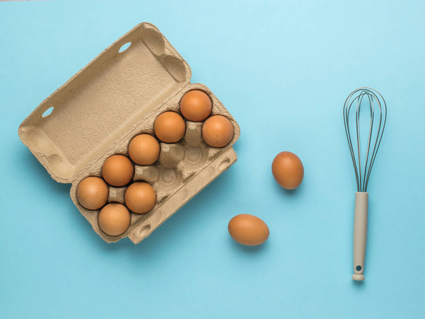 An open box of eggs and a whisk on a blue background. Natural products and kitchen equipment. - Fotoğraf, Görsel