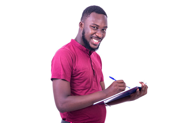 retrato de joven feliz guapo usando camiseta roja, escribiendo en cuaderno con pluma mientras sonríe a la cámara. - Foto, imagen