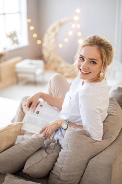 Hermosa mujer joven sensual con el pelo rubio con una camisa blanca libro de lectura en el sofá en el hogar moderno - Foto, imagen