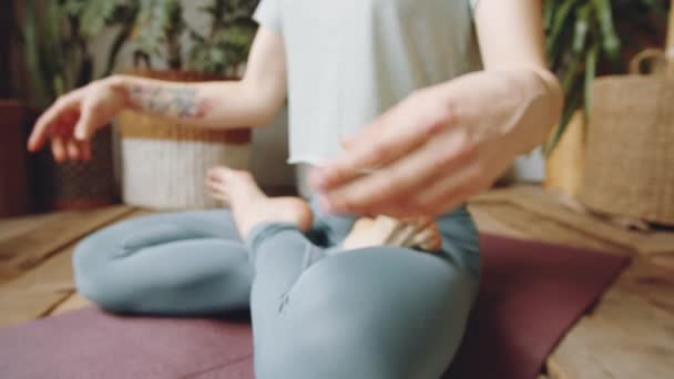 Primer plano plano de mujer descalza irreconocible en ropa deportiva sentada en pose de loto sobre esterilla y cogida de la mano en mudras durante la práctica de yoga en casa - Imágenes, Vídeo