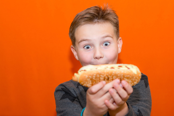 Έκπληκτος 10s αγόρι θέλει να δαγκώσει ένα μεγάλο hot dog.Closeup πορτοκαλί στούντιο σε εσωτερικούς χώρους.Σώμα θετικό. - Φωτογραφία, εικόνα