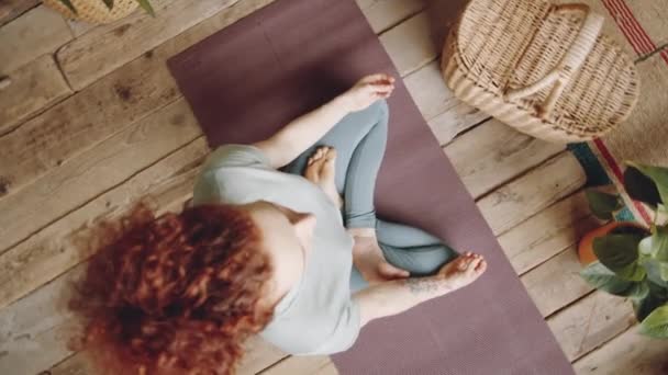 Ampliar vista superior plano de mujer descalza con pelo rizado rojo sentada en pose de loto sobre estera y cogida de la mano en mudras mientras practica meditación de yoga en casa en habitación con suelo de madera - Imágenes, Vídeo