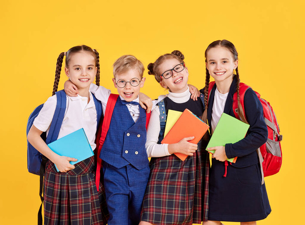 Optimistische Grundschüler in Schuluniform mit Rucksäcken blicken vor gelbem Hintergrund in die Kamera - Foto, Bild