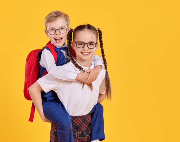 Cheerful girl in school uniform and glasses giving piggyback ride to happy little brother on yellow background - Photo, Image