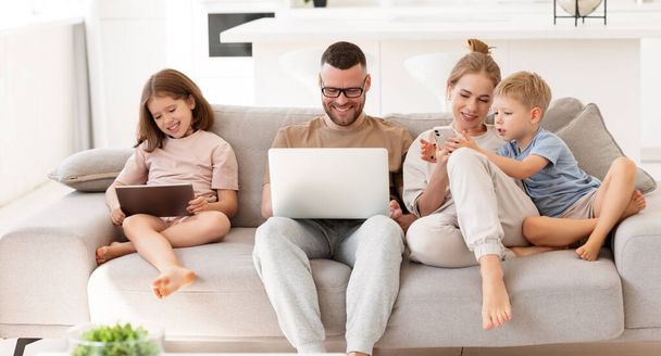 Junge glückliche Familien, Eltern mit zwei Kindern, die moderne Technologien nutzen, während sie Zeit miteinander zu Hause verbringen. Vater, Mutter und Kinder mit Laptop, digitalem Tablet und Smartphone entspannt auf dem Sofa - Foto, Bild