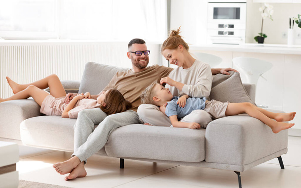 Parents expressing love to kids. Beautiful happy family of four, mother, father and two cute children spending time together at home, embracing and relaxing on sofa in living room - Foto, Imagen
