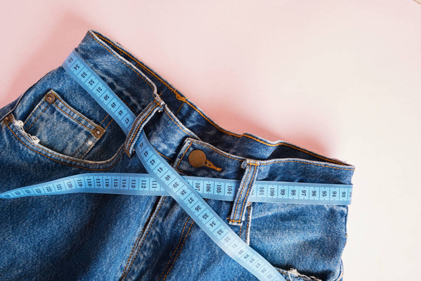 blue measuring tape is threaded into jeans instead of a belt, blue jeans on a pink background, copy space, weight loss and weight control concept, measuring body volume - Foto, immagini