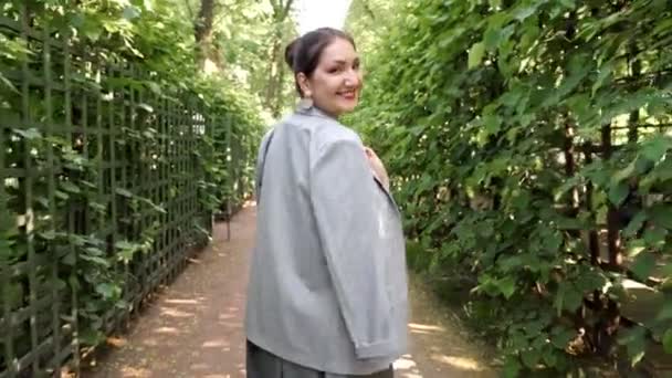 Mujer joven camina por el jardín y se da la vuelta sonriendo - Imágenes, Vídeo