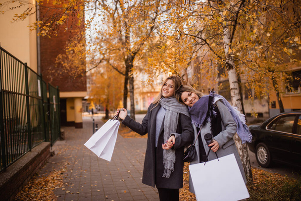 Zwei hübsche blonde Frauen stehen im Park aneinander gelehnt und posieren mit Taschen in den Händen. Herbsteinkauf - Foto, Bild