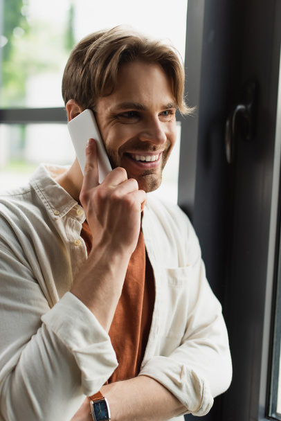 uśmiechnięty młody mężczyzna w beżowej koszuli rozmawiający przez telefon komórkowy w nowoczesnym biurze - Zdjęcie, obraz