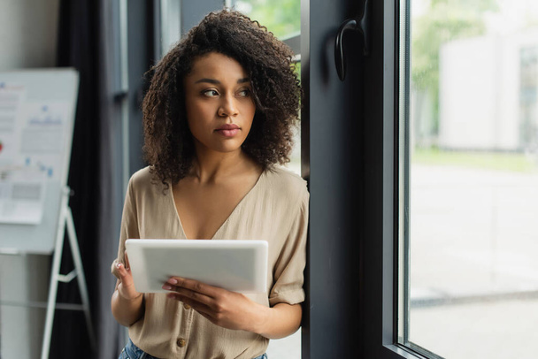 thoughtful african american woman standing near window with digital tablet in hand in modern office - Photo, Image