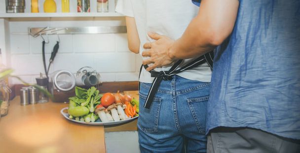 Pareja vista trasera abrazó amorosamente su cintura mientras se preparaba para cocinar y verduras frescas en la cocina. - Foto, imagen