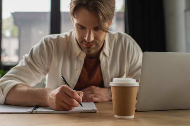 konzentrierter junger Mann sitzt am Schreibtisch neben Pappbecher und Laptop und schreibt mit Stift in Notizbuch in modernem Büro - Foto, Bild