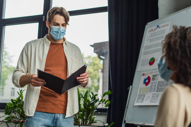 young man in medical mask holding folder with papers near flip chart and african american colleague in office - 写真・画像