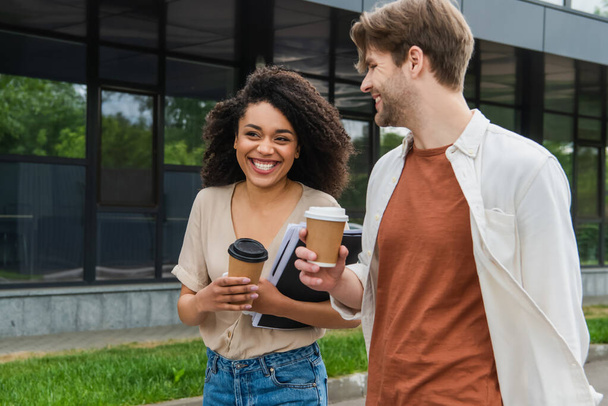 sourire jeune couple interracial avec des tasses à café en papier et dossier avec des documents marchant sur la rue près du bâtiment moderne - Photo, image