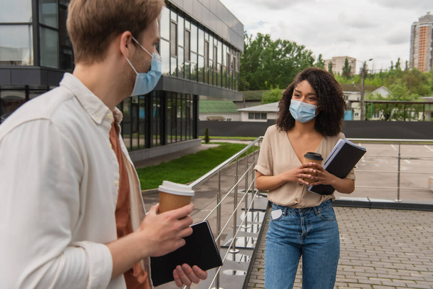 giovane coppia interrazziale in maschere mediche parlando in strada con il caffè per andare vicino edificio di vetro - Foto, immagini