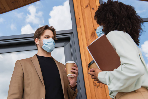 low angle view of young interracial colleagues in medical masks talking with paper cups in hands near building - Foto, Imagem