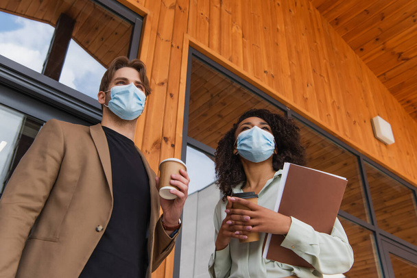 low angle view of young interracial colleagues in medical masks standing with paper cups in hands on street near building - Photo, Image