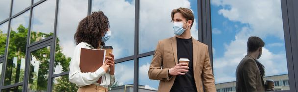 young interracial colleagues in medical masks walking with paper cups and documents near glass building, banner - Photo, Image