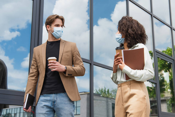 young interracial colleagues in medical masks walking with coffee paper cups and documents near modern glass building - Fotó, kép