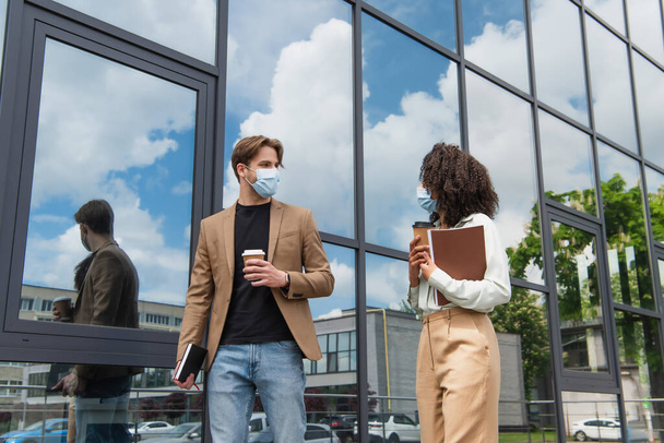 young interracial colleagues in medical masks walking with coffee paper cups and documents near modern building - Photo, Image