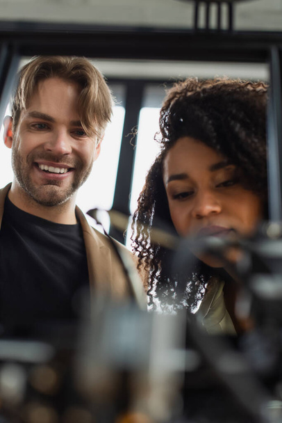 alegres jóvenes colegas interracial mirando borrosa impresora 3D en la oficina moderna - Foto, imagen