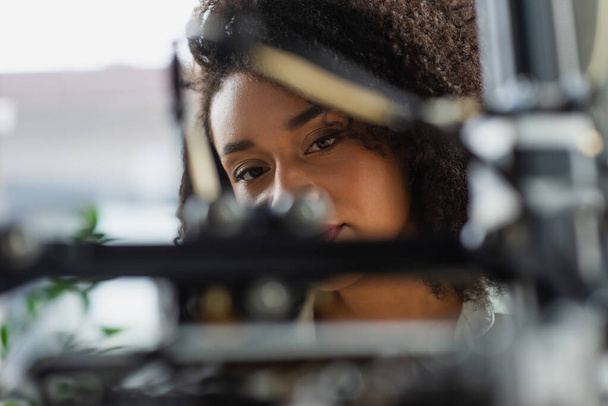 vista de cerca de la mujer afroamericana mirando borrosa impresora 3D en la oficina - Foto, imagen