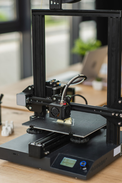 Imprimante 3D créant le modèle en plastique près des figures floues et ordinateur portable dans le bureau moderne - Photo, image