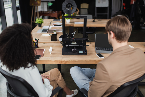 vista trasera de los jóvenes diseñadores interracial sentados cerca de la impresora 3D en el espacio abierto moderno - Foto, Imagen