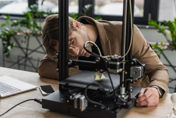 concentrado joven diseñador mirando a la impresora 3D produciendo figura de plástico cerca de la computadora portátil y el teléfono celular en la oficina moderna - Foto, imagen