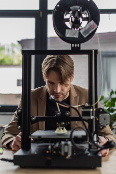 enfocado joven diseñador mirando a la impresora 3D producir modelo en la oficina moderna - Foto, Imagen