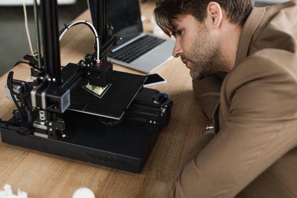 keskittynyt nuori mies etsii 3D-tulostin, jossa on muovinen luku modernissa toimistossa - Valokuva, kuva