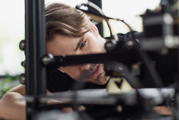 στενή άποψη του νεαρού σχεδιαστή που εξετάζει την εργασία 3D εκτυπωτή στο γραφείο - Φωτογραφία, εικόνα