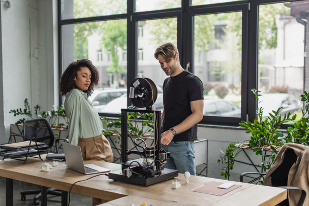 colegas interracial de pie cerca de la impresora 3D, ordenador portátil y figuras de plástico en la oficina moderna - Foto, imagen