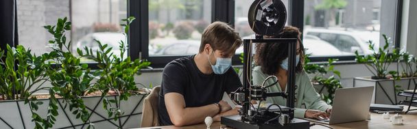 interracial colleagues in medical masks working with laptop near 3D printer in modern office, banner - Foto, imagen