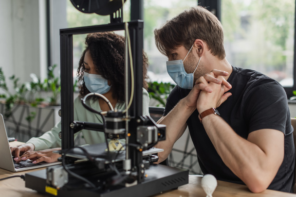 межрасовые коллеги в медицинских масках, работающие с 3D принтером и ноутбуком в современном офисе - Фото, изображение