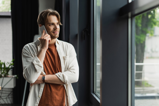 positive young man in beige shirt talking on cellphone near window in modern office - Fotoğraf, Görsel