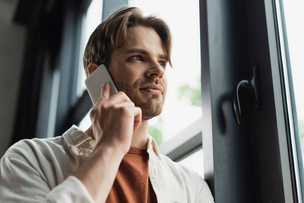 positiivinen nuori mies beige paita puhuu kännykkään ikkunan lähellä modernissa toimistossa - Valokuva, kuva