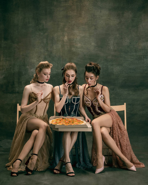 Pretty women in vintage 20s dresses. Young girls in art action sitting together and posing on wall background. - Foto, afbeelding