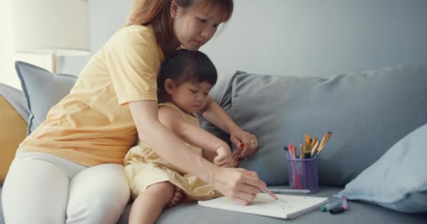 Neşeli Asya ailesi anneleri kızlara boya kullanım albümü ve renkli kalemler öğretiyor. Oturma odasındaki kanepede rahatça eğleniyorlar. Birlikte zaman geçirmek, sosyal uzaklık, koronavirüs karantinası.. - Video, Çekim