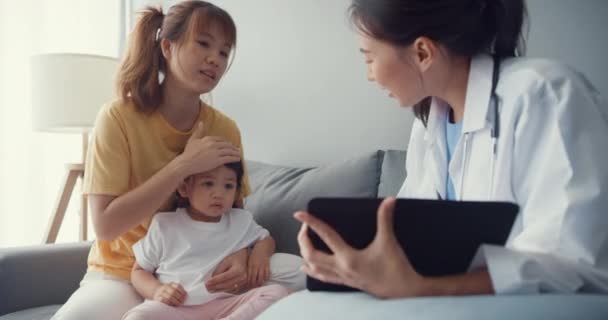 Mladá Asie ženské pediatr lékař a malá holka pacient pomocí digitální tablet sdílení dobré zprávy o zdravotním testu s šťastný maminka sedět na gauči v domě. Zdravotní pojištění, Návštěva pacienta doma koncepce. - Záběry, video