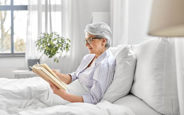 old woman in glasses reading book in bed at home - Фото, зображення