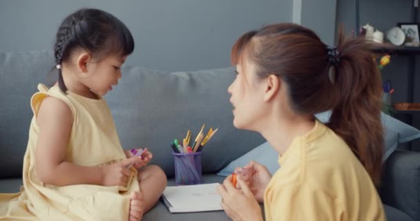 Счастливая веселая азиатская семья мама учить девочек краски использовать альбом и красочные карандаши весело отдохнуть на диване в гостиной в доме. Проводим время вместе, социальная дистанция, карантин для коронавируса. - Кадры, видео
