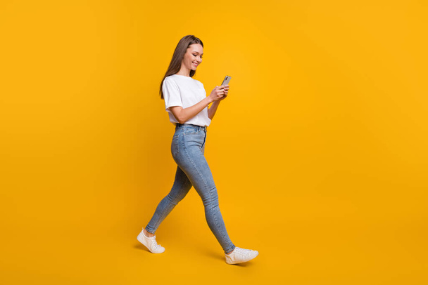 Teljes hossz testméret fotó női blogger lépésről előre gépelés üzenet mobiltelefon elszigetelt élénk sárga színű háttér - Fotó, kép