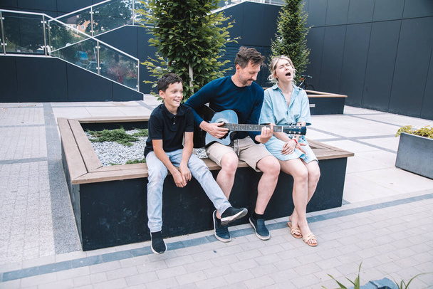 Familia joven canta y toca la guitarra en el día de verano en la plaza de la ciudad. El concepto de familia feliz. Estilo de vida.  - Foto, Imagen