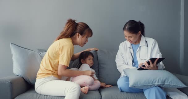 Jovem asiática pediatra e pequena paciente usando tablet digital compartilhando boas notícias de teste de saúde com a mãe feliz sentar no sofá em casa. Seguro médico, Visite o conceito de paciente em casa. - Filmagem, Vídeo