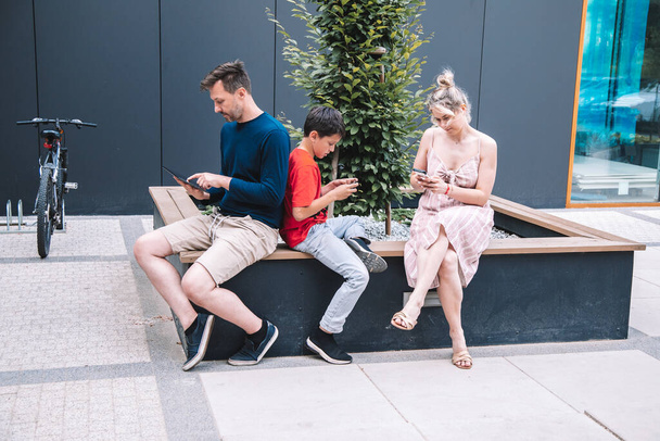 Młodzi rodzice i syn korzystają z technologii bezprzewodowej podczas relaksu na placu miasta w letni dzień. Wysokiej jakości zdjęcie. Styl życia - Zdjęcie, obraz