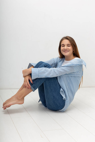 young caucasian pretty girl with long hair in shirt, blue jeans sits at studio - Zdjęcie, obraz