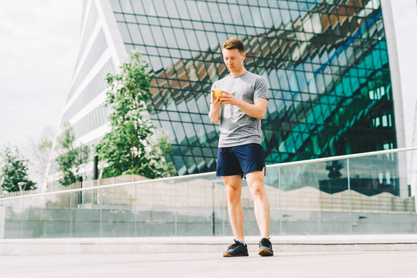 Tired Young man runner standing and using mobile phone and relaxing after sport training. Holding water bottle while doing fitness workout in city urban street, cloudy sky at summer. - Fotografie, Obrázek
